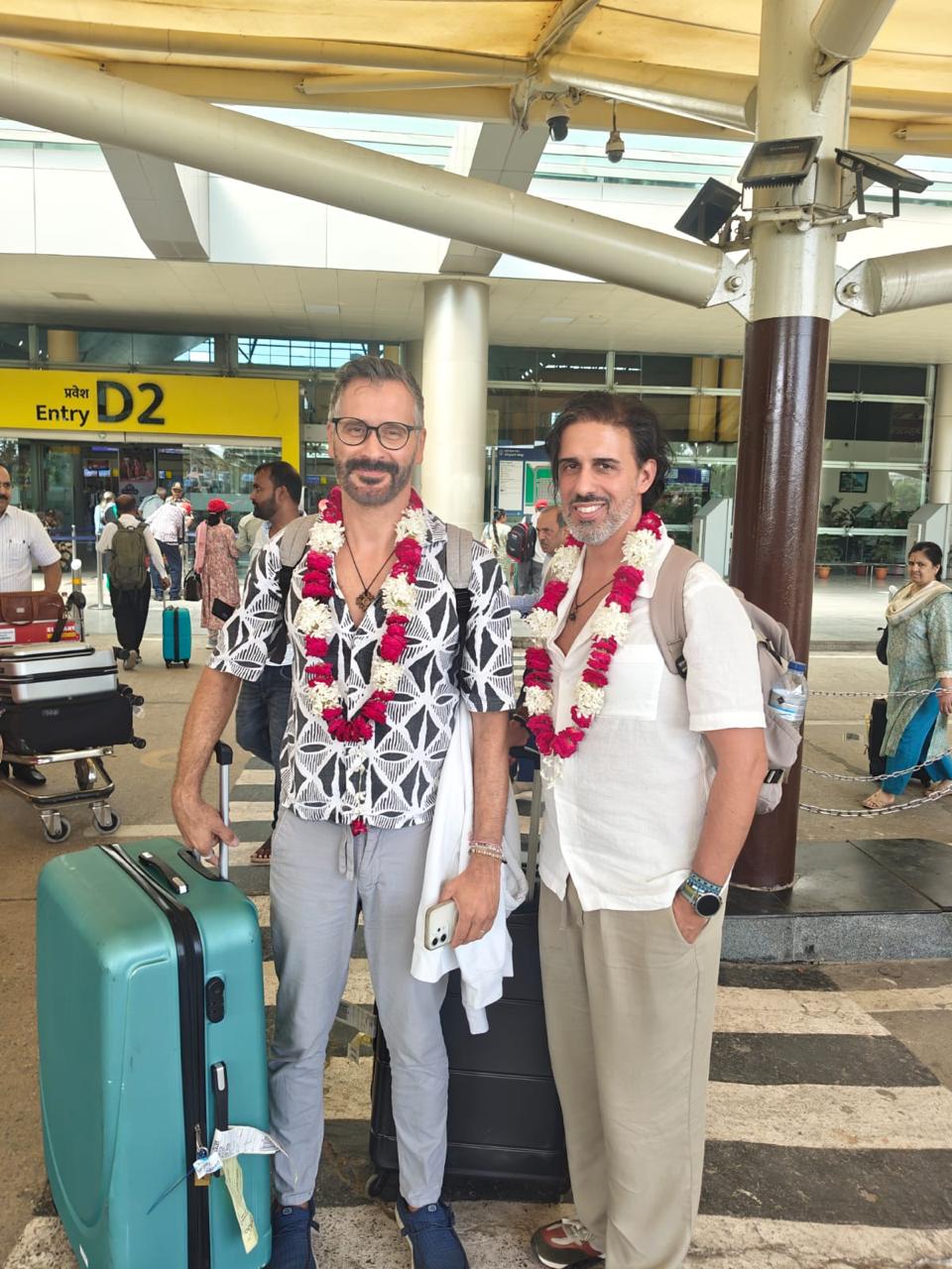 Two travellers with flower garlands and luggage outside the airport