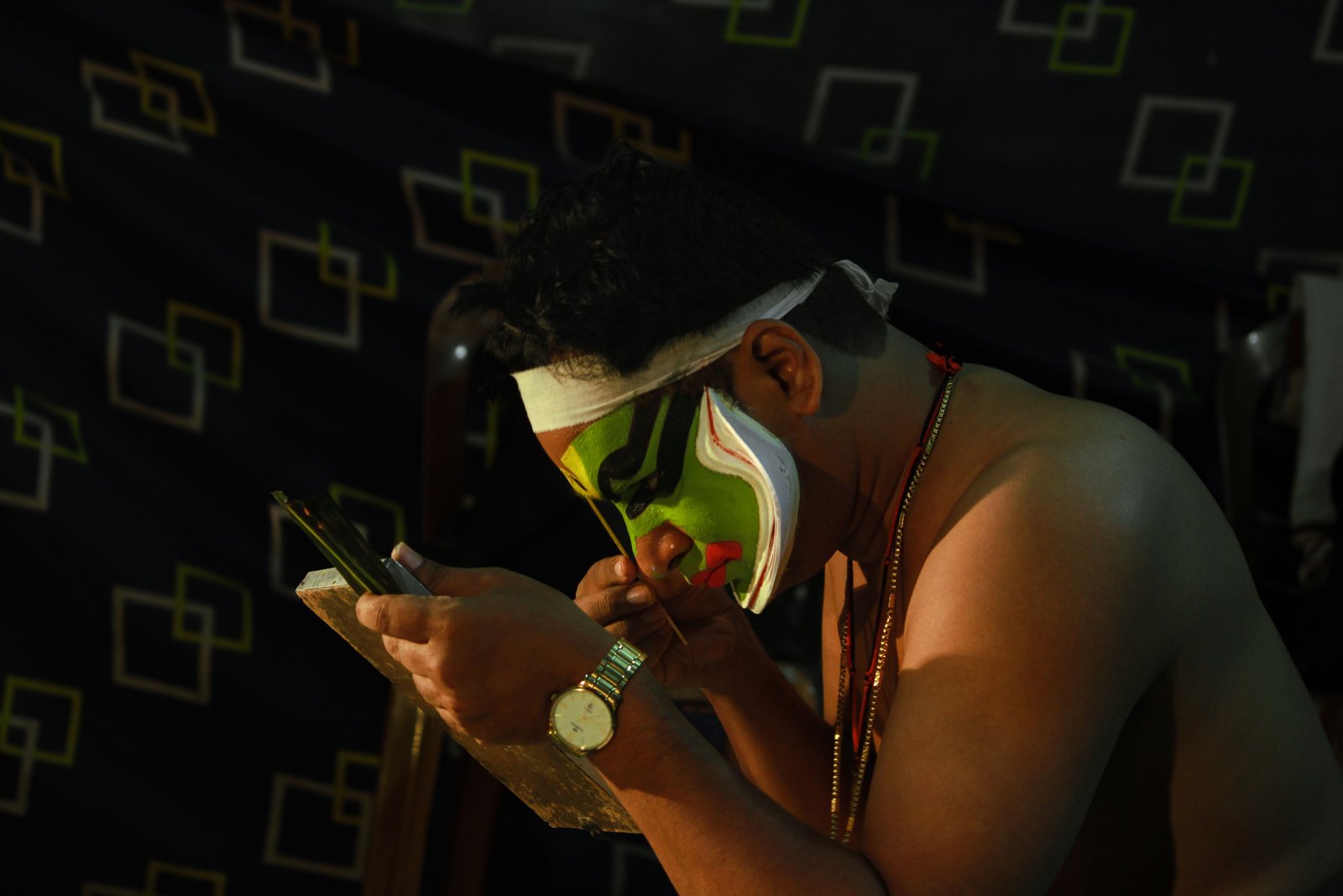 A Kathakali performer applying make-up while looking into a small mirror.